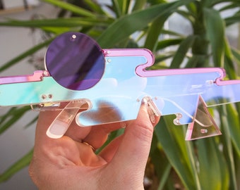 Peut inclure: Une paire de lunettes de soleil iridescentes rose et bleu avec une lentille ronde violette. Les lunettes sont en plastique et ont un design géométrique unique.