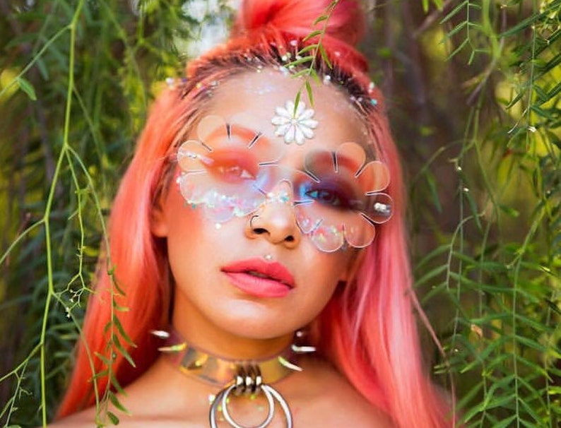 May include: A person with pink hair wearing clear flower-shaped sunglasses with iridescent glitter accents. The sunglasses have a white flower design on the bridge.