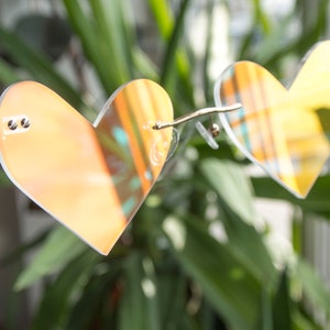 May include: Heart-shaped sunglasses with mirrored lenses in a warm orange hue. The frame is clear plastic with gold accents.