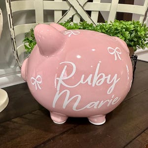 May include: A pink ceramic piggy bank with the name "Ruby Marie" in white script. The bank has a bow design and is sitting on a wooden surface. The piggy bank has four small feet.