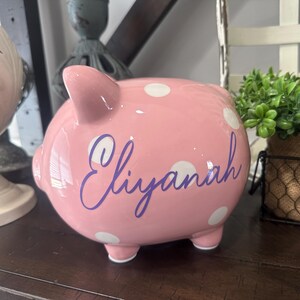 May include: A pink ceramic piggy bank with white polka dots and the name "Eliyanah" in purple script. The piggy bank has four small feet and a slot on top for inserting coins. It is a decorative item.