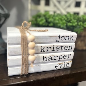 May include: A stack of four white wooden books tied with twine and wooden beads. Each book is labeled with a name in black font: Josh, Kristen, Harper, and Evie. The books are on a dark brown surface.