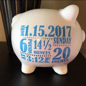 Peut inclure: Tirelire en c&eacute;ramique blanche avec texte bleu. Le texte comprend la date 1.15.2017, le poids 2,7 kg, la longueur 50,8 cm et l'heure 15h12. La tirelire a quatre petits pieds et une fente sur le dessus pour ins&eacute;rer des pi&egrave;ces.