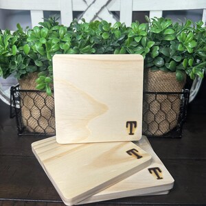 Puede incluir: Una pila de cuatro posavasos cuadrados de madera. Cada posavasos es de color madera natural claro con un borde redondeado. La letra "T" est&aacute; grabada en la esquina inferior derecha de cada posavasos. Los posavasos est&aacute;n apilados sobre una superficie de madera oscura.