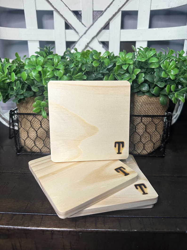 Puede incluir: Una pila de cuatro posavasos cuadrados de madera. Cada posavasos es de color madera natural claro con un borde redondeado. La letra "T" est&aacute; grabada en la esquina inferior derecha de cada posavasos. Los posavasos est&aacute;n apilados sobre una superficie de madera oscura.
