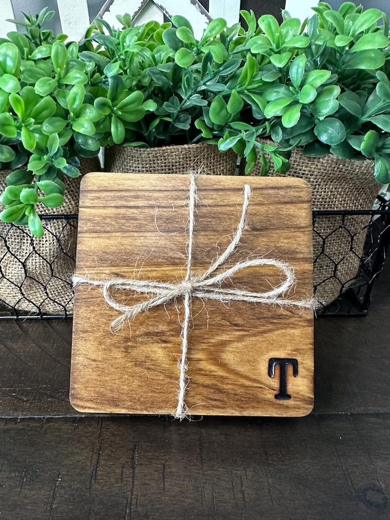 Puede incluir: Un posavasos cuadrado de madera con tinte oscuro, atado con hilo de bramante. El posavasos tiene una letra negra "T" en la esquina inferior derecha. Plantas verdes en macetas forradas de arpillera en el fondo.