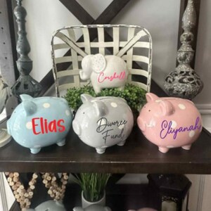 Peut inclure: Une collection de tirelires en c&eacute;ramique de diff&eacute;rentes couleurs, dont bleu, gris et rose, chacune avec des pois blancs. Les tirelires sont personnalis&eacute;es avec des noms et des phrases dans diff&eacute;rentes polices. Un cochon tirelire en forme d'&eacute;l&eacute;phant est &eacute;galement pr&eacute;sent.