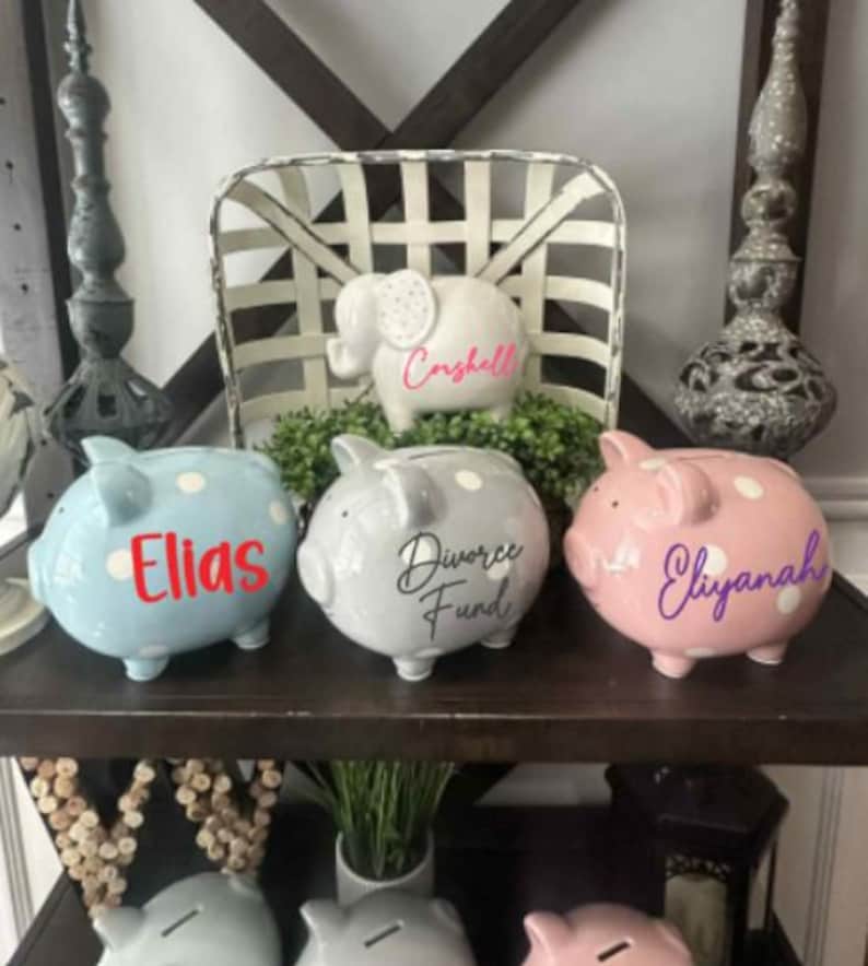 Peut inclure: Une collection de tirelires en c&eacute;ramique de diff&eacute;rentes couleurs, dont bleu, gris et rose, chacune avec des pois blancs. Les tirelires sont personnalis&eacute;es avec des noms et des phrases dans diff&eacute;rentes polices. Un cochon tirelire en forme d'&eacute;l&eacute;phant est &eacute;galement pr&eacute;sent.