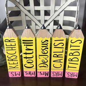 May include: Five wooden pencil-shaped blocks with yellow bodies, natural wood tips, and pink erasers. Each block features black text reading MS. KERSCHER, MRS. COTTRILL, MRS. DE JESUS, MRS. CARLISI, and MRS. TIBBITS printed vertically on the yellow section.