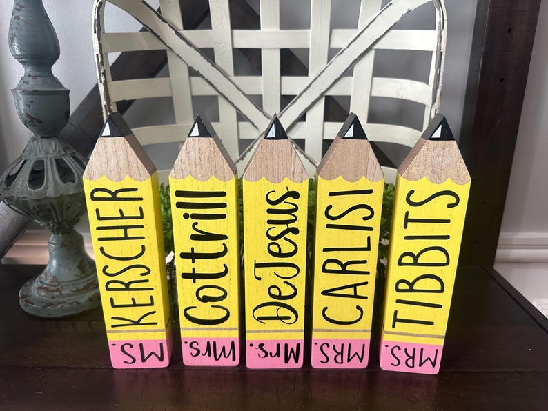 May include: Five wooden pencil-shaped blocks with yellow bodies, natural wood tips, and pink erasers. Each block features black text reading MS. KERSCHER, MRS. COTTRILL, MRS. DE JESUS, MRS. CARLISI, and MRS. TIBBITS printed vertically on the yellow section.