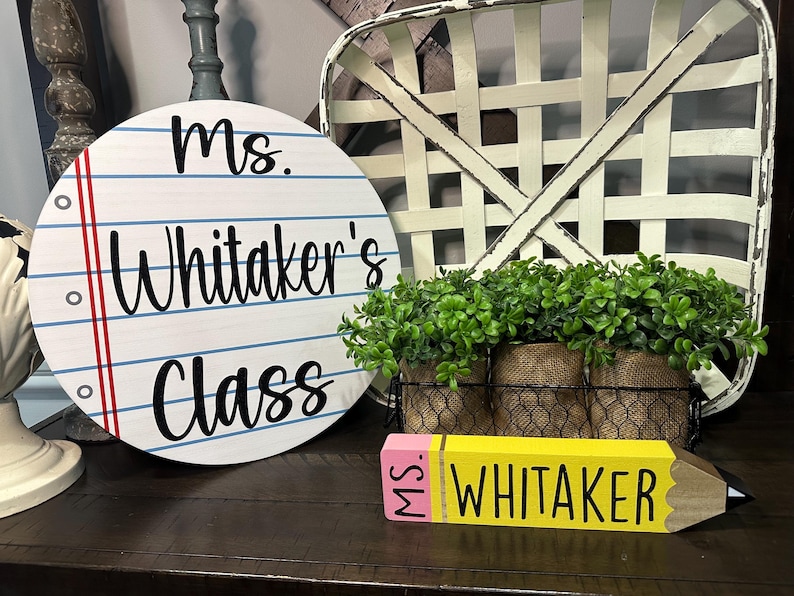 May include: A round wooden sign featuring a white background with blue horizontal lines and a red vertical margin line, printed with the black text Ms. Whitaker's Class. Beside it is a yellow pencil-shaped wooden block labeled Ms. Whitaker.