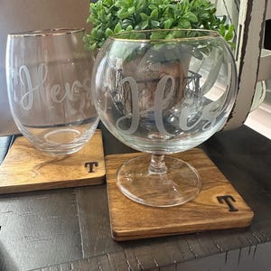 Puede incluir: Dos copas de vino transparentes con bordes dorados, una con la palabra "Hers" grabada y la otra con "His". Se apoyan en posavasos cuadrados de madera con la letra "T".