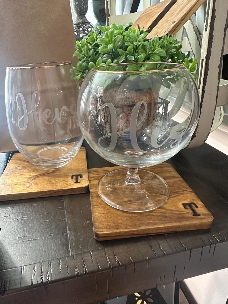 Puede incluir: Dos copas de vino transparentes con bordes dorados, una con la palabra "Hers" grabada y la otra con "His". Se apoyan en posavasos cuadrados de madera con la letra "T".