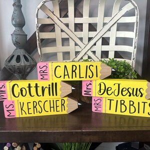 May include: Five yellow wooden pencil-shaped blocks with pink erasers and black text. The blocks are personalized with the names Mrs. Carlisi, Mrs. Cottrill, Ms. Kerscher, Mrs. DeJesus, and Mrs. Tibbits printed in black script on the yellow surface.