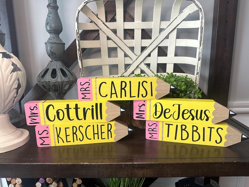 May include: Five yellow wooden pencil-shaped blocks with pink erasers and black text. The blocks are personalized with the names Mrs. Carlisi, Mrs. Cottrill, Ms. Kerscher, Mrs. DeJesus, and Mrs. Tibbits printed in black script on the yellow surface.