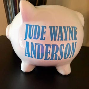 Peut inclure: Une tirelire en c&eacute;ramique blanche avec le nom "JUDE WAYNE ANDERSON" en lettres bleues. La tirelire a la forme classique d'un cochon avec quatre petites pattes, une queue et une fente sur le dessus pour ins&eacute;rer des pi&egrave;ces. Le cochon est pos&eacute; sur une surface sombre.