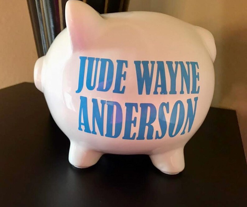 Peut inclure: Une tirelire en c&eacute;ramique blanche avec le nom "JUDE WAYNE ANDERSON" en lettres bleues. La tirelire a la forme classique d'un cochon avec quatre petites pattes, une queue et une fente sur le dessus pour ins&eacute;rer des pi&egrave;ces. Le cochon est pos&eacute; sur une surface sombre.
