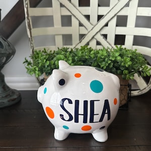 May include: White ceramic piggy bank with the name "SHEA" in navy blue. The bank is decorated with orange, teal, and navy blue polka dots. The piggy bank is on a dark brown surface.