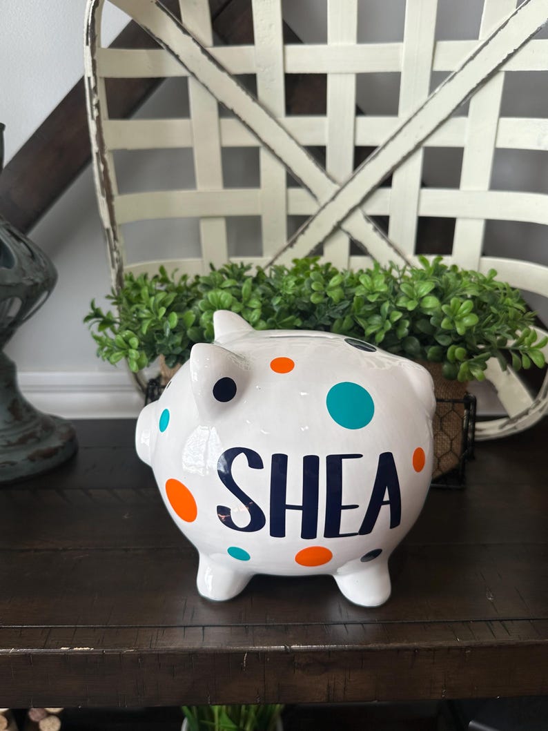May include: White ceramic piggy bank with the name "SHEA" in navy blue. The bank is decorated with orange, teal, and navy blue polka dots. The piggy bank is on a dark brown surface.