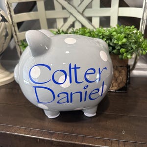 May include: A gray ceramic piggy bank with white polka dots and the name "Colter Daniel" in blue script. The bank has four small feet and a slot on top for inserting coins. A decorative item for a child's room.