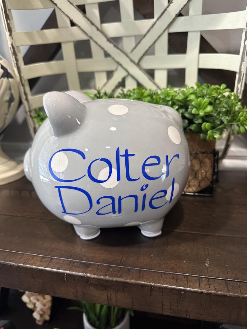 May include: A gray ceramic piggy bank with white polka dots and the name "Colter Daniel" in blue script. The bank has four small feet and a slot on top for inserting coins. A decorative item for a child's room.