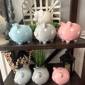 May include: A collection of ceramic piggy banks in various colors, including blue, gray, and pink, each adorned with white polka dots. The banks are displayed on a dark wooden shelf, alongside a white elephant figurine and decorative items.