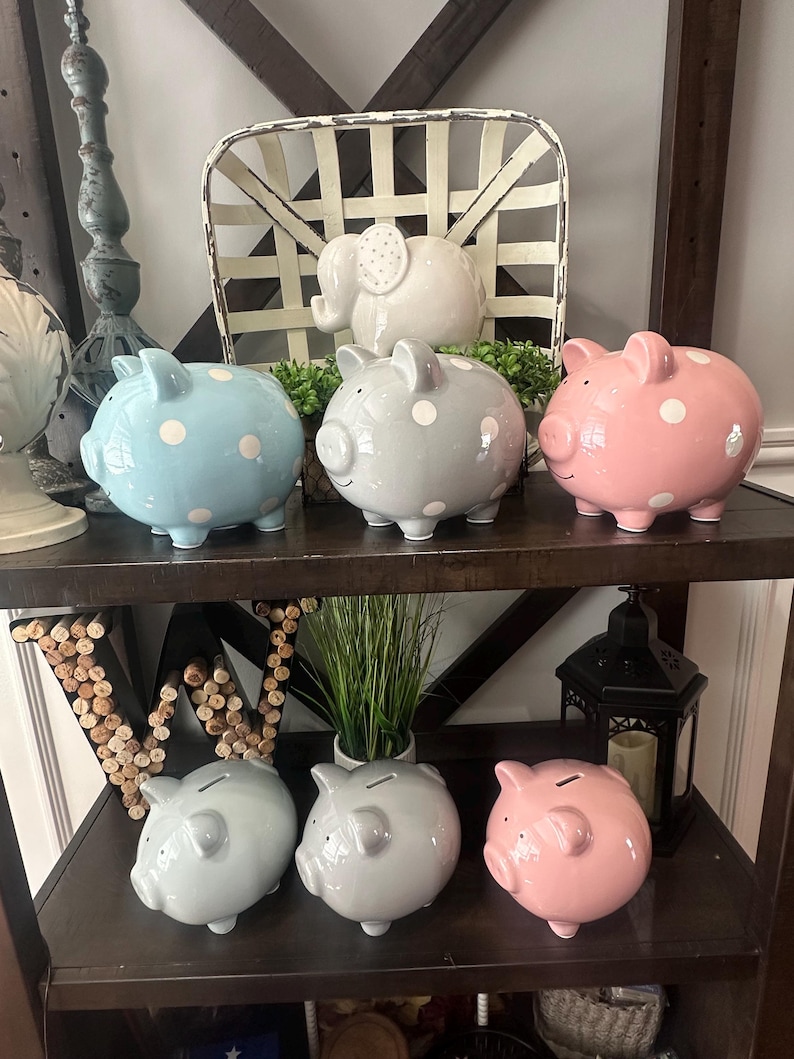 May include: A collection of ceramic piggy banks in various colors, including blue, gray, and pink, each adorned with white polka dots. The banks are displayed on a dark wooden shelf, alongside a white elephant figurine and decorative items.