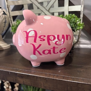 May include: A pink ceramic piggy bank with white polka dots. The name "Aspyn Kate" is written in pink script across the front. The piggy bank is sitting on a wooden shelf.