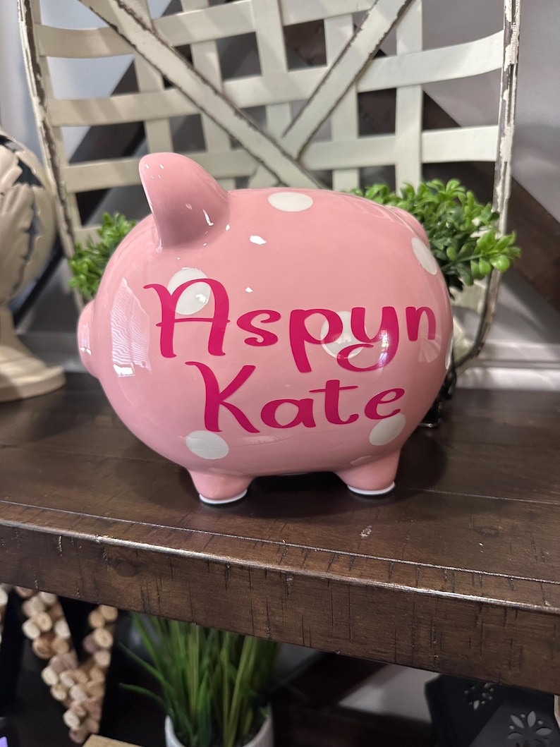 May include: A pink ceramic piggy bank with white polka dots. The name "Aspyn Kate" is written in pink script across the front. The piggy bank is sitting on a wooden shelf.