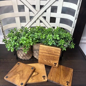 Puede incluir: Cuatro posavasos cuadrados de madera, cada uno con la letra "T" en negro. Los posavasos son de varios tonos marrones y est&aacute;n sobre una superficie de madera oscura. Una cesta con vegetaci&oacute;n est&aacute; en el fondo.