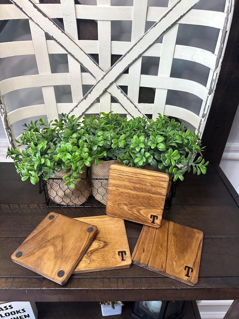 Puede incluir: Cuatro posavasos cuadrados de madera, cada uno con la letra "T" en negro. Los posavasos son de varios tonos marrones y est&aacute;n sobre una superficie de madera oscura. Una cesta con vegetaci&oacute;n est&aacute; en el fondo.