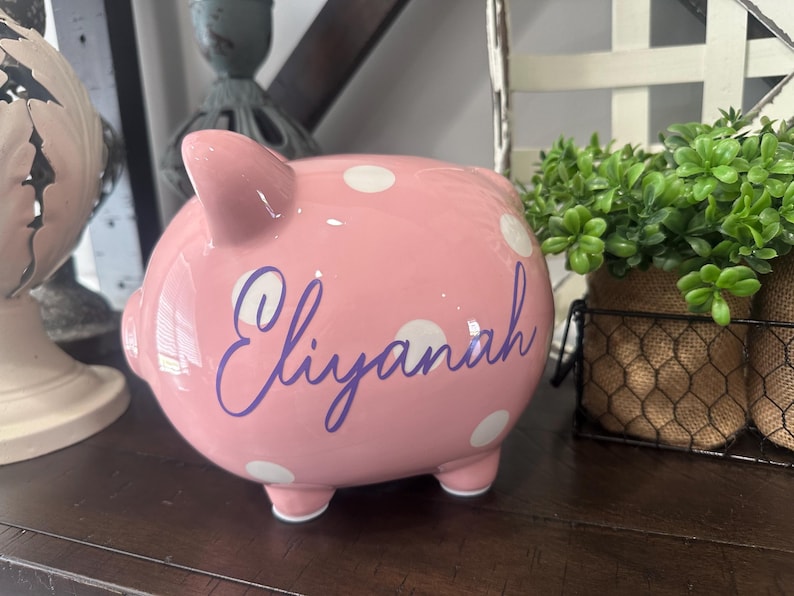 May include: A pink ceramic piggy bank with white polka dots and the name "Eliyanah" in purple script. The piggy bank has four small feet and a slot on top for inserting coins. It is a decorative item.