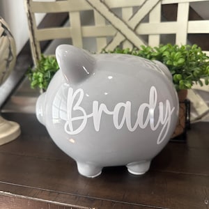 May include: A gray ceramic piggy bank with the name "Brady" in white script. The bank has four small feet and a slot on top for inserting coins. The piggy bank is a classic design, perfect for saving money.
