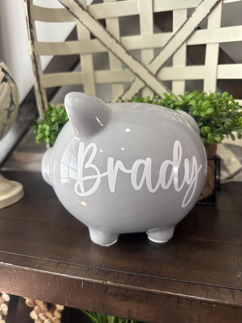 May include: A gray ceramic piggy bank with the name "Brady" in white script. The bank has four small feet and a slot on top for inserting coins. The piggy bank is a classic design, perfect for saving money.