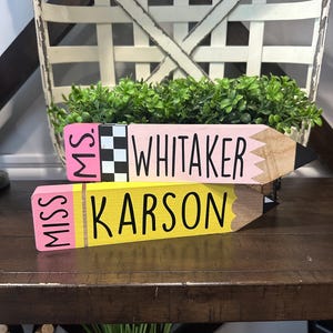 May include: Two wooden pencil-shaped signs with flat ends. The top sign is pink with a black and white checkered band and the text "MS. WHITAKER". The bottom sign is yellow with the text "MISS KARSON". Both have pointed, dark-tipped ends.