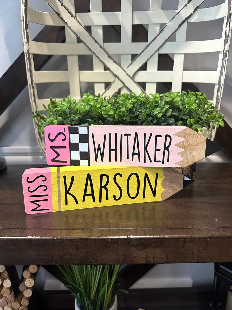 May include: Two wooden pencil-shaped signs with flat ends. The top sign is pink with a black and white checkered band and the text "MS. WHITAKER". The bottom sign is yellow with the text "MISS KARSON". Both have pointed, dark-tipped ends.