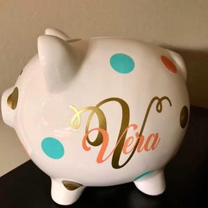 May include: A white ceramic piggy bank with teal, orange, and gold polka dots. The name "Vera" is written in gold and coral script. The piggy bank has four small feet and two ears.