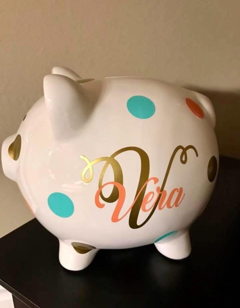 May include: A white ceramic piggy bank with teal, orange, and gold polka dots. The name "Vera" is written in gold and coral script. The piggy bank has four small feet and two ears.