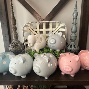 Peut inclure: Une collection de tirelires en c&eacute;ramique de diff&eacute;rentes couleurs, dont bleu, gris et rose, certaines avec des pois blancs. Une tirelire en forme d'&eacute;l&eacute;phant et des objets d&eacute;coratifs sont &eacute;galement pr&eacute;sents.