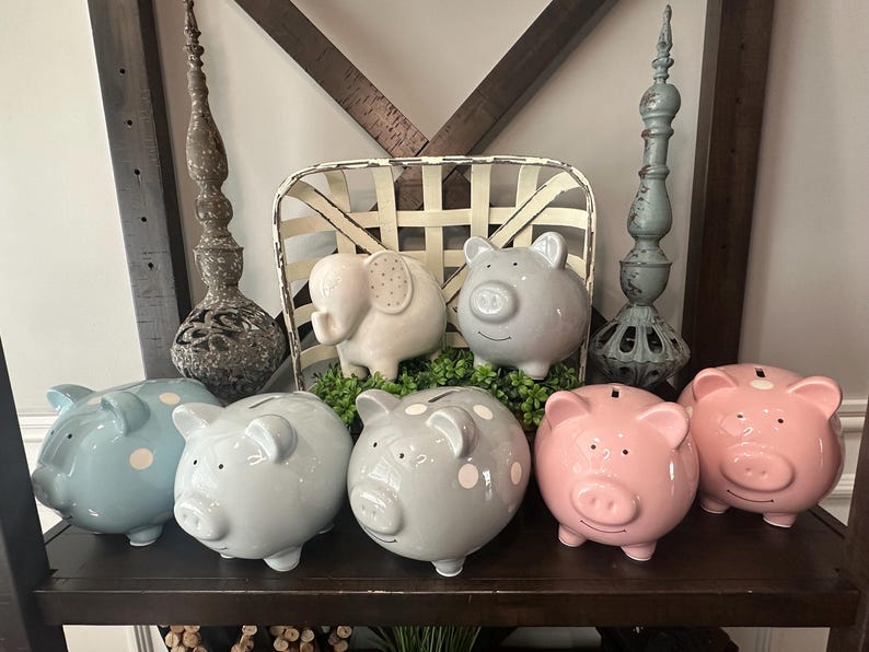 Peut inclure: Une collection de tirelires en c&eacute;ramique de diff&eacute;rentes couleurs, dont bleu, gris et rose, certaines avec des pois blancs. Une tirelire en forme d'&eacute;l&eacute;phant et des objets d&eacute;coratifs sont &eacute;galement pr&eacute;sents.