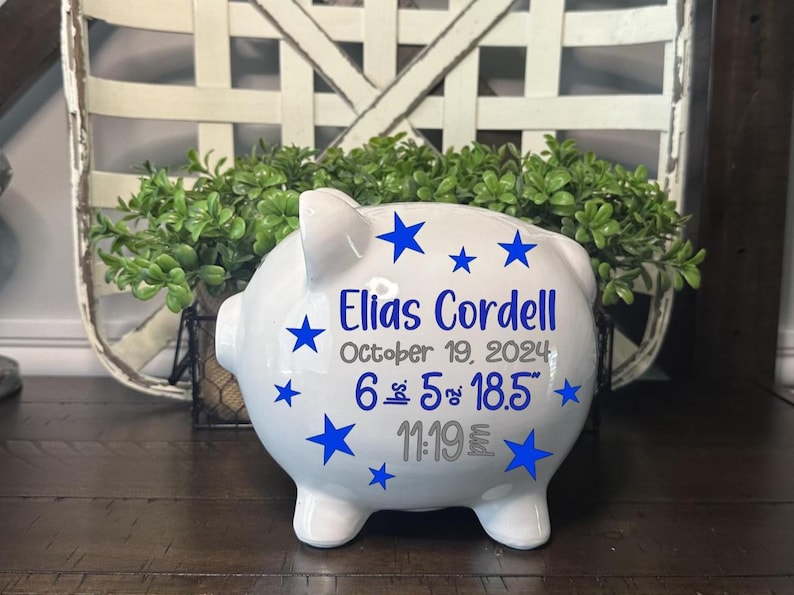 May include: White ceramic piggy bank with blue star accents and the name "Elias Cordell" in blue. The text includes the date "October 19, 2024", and measurements of 6 lbs 5 oz and 18.5 inches, and the time 11:19.