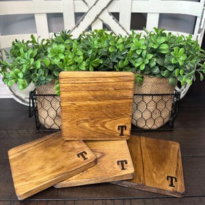 Puede incluir: Juego de cuatro posavasos cuadrados de madera con tinte oscuro y la letra "T" grabada en cada uno. Los posavasos est&aacute;n dispuestos sobre una superficie de madera oscura, con una cesta decorativa y vegetaci&oacute;n en el fondo.