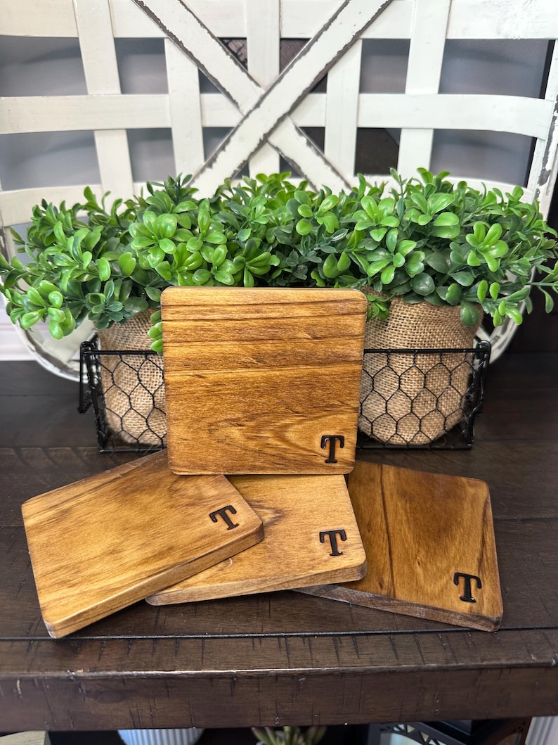 Puede incluir: Juego de cuatro posavasos cuadrados de madera con tinte oscuro y la letra "T" grabada en cada uno. Los posavasos est&aacute;n dispuestos sobre una superficie de madera oscura, con una cesta decorativa y vegetaci&oacute;n en el fondo.
