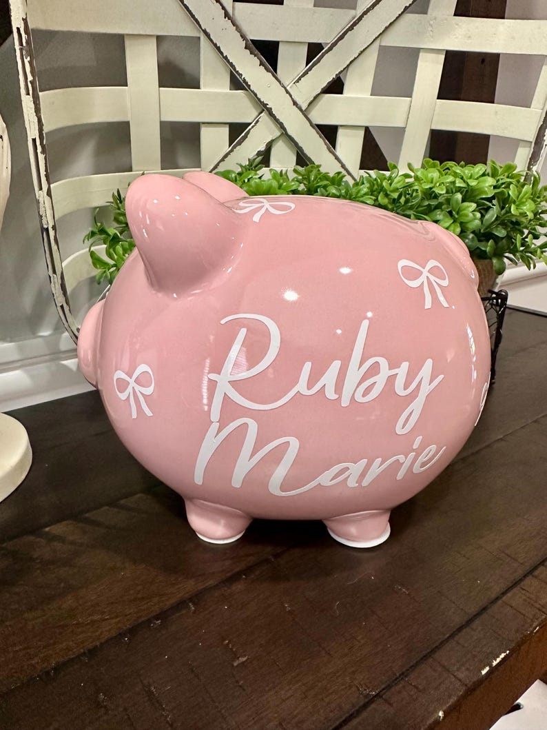 May include: A pink ceramic piggy bank with the name "Ruby Marie" in white script. The bank has a bow design and is sitting on a wooden surface. The piggy bank has four small feet.