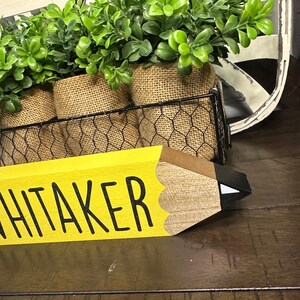 May include: A wooden pencil-shaped sign painted yellow with the name WHITAKER in black capital letters. The pencil features a brown wooden tip and a black lead point with a white accent. Behind the sign is a black wire basket holding three burlap-wrapped pots containing artificial green leafy plants.