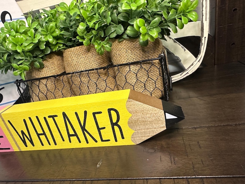 May include: A wooden pencil-shaped sign painted yellow with the name WHITAKER in black capital letters. The pencil features a brown wooden tip and a black lead point with a white accent. Behind the sign is a black wire basket holding three burlap-wrapped pots containing artificial green leafy plants.