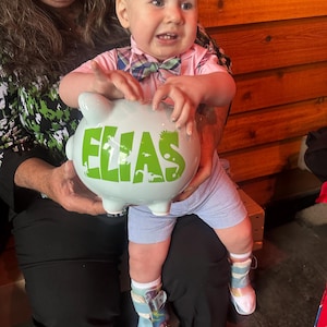 May include: A light blue piggy bank with the name "ELIAS" in green letters is held by a baby. The baby is wearing a pink shirt, blue shorts, and colorful socks. The baby is being held by a person.