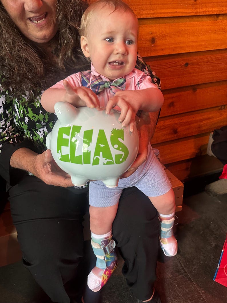 May include: A light blue piggy bank with the name "ELIAS" in green letters is held by a baby. The baby is wearing a pink shirt, blue shorts, and colorful socks. The baby is being held by a person.