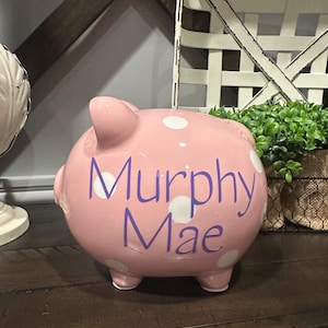 May include: A pink ceramic piggy bank with white polka dots and the name "Murphy Mae" in purple script. The bank has four small feet and a slot on top for inserting coins. A decorative item for a child's room.