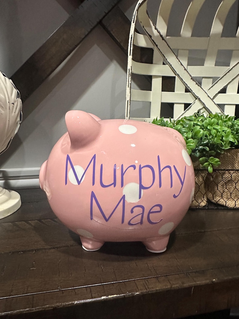May include: A pink ceramic piggy bank with white polka dots and the name "Murphy Mae" in purple script. The bank has four small feet and a slot on top for inserting coins. A decorative item for a child's room.